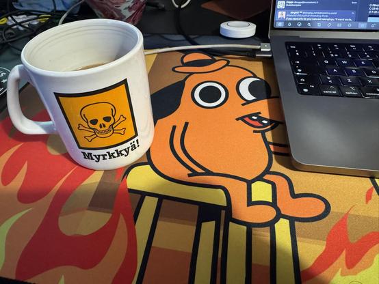 Valkoinen kahvimuki, jossa on keltainen varoitusmerkki ja teksti "Myrkkyä!", pöydällä värikkäällä hiirimatolla, jossa on "This is fine" -koiran hahmo tulen keskellä. Muki on osittain täynnä kahvia ja vieressä on kannettavan tietokoneen kulma.