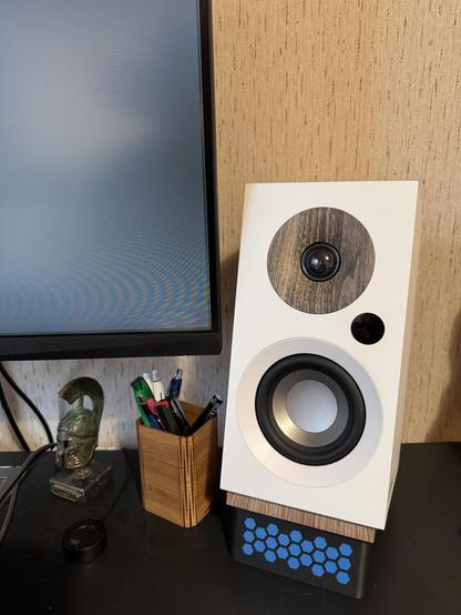 Työpöydällä tietokonenäytön vieressä oleva valkoinen kaiutin, jonka alla on sinisillä hunajakennokuvioilla koristeltu teline; vieressä kynäpurkki ja pieni koristepatsas.