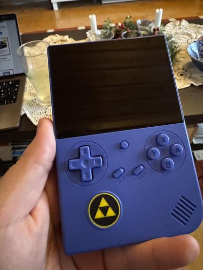 Henkilö pitää kädessään sinistä retrohenkistä käsikonsolia, jossa on suuri musta näyttö, ristinappaimet, A–B–X–Y -painikkeet sekä Triforce-symboli etupuolella.