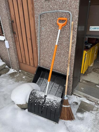 Lumella on lunta, jossa on talviset työvälineet: lumikola ja pienipuhuri, valkoinen kivi ja taustalla rakennuksen seinä.