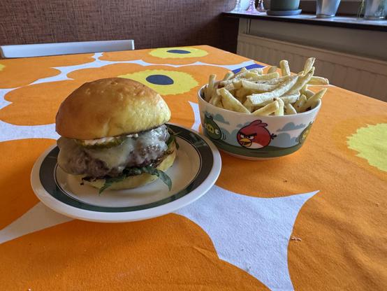 Hampurilainen ja kulhollinen ranskalaisia perunoita pöydällä, jonka pöytäliina on oranssi ja sisältää kukkakuvioita.