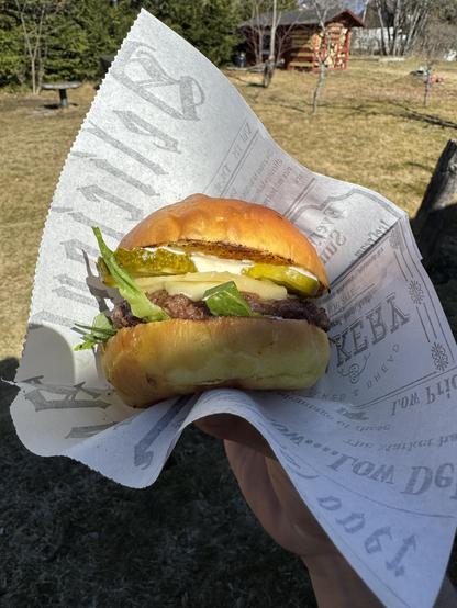 Hampurilainen kädessä, ulkona puutarhassa, taustalla puita ja pieni rakennus.