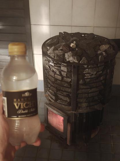 Etualalla kädessä pieni musta Vichy pullo. Taka alalla puukiuas jossa vielä hiillos. Hieman huuruinen kuva.